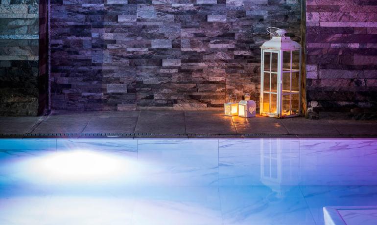 Piscina illuminata con candele e orologio su parete di pietra.