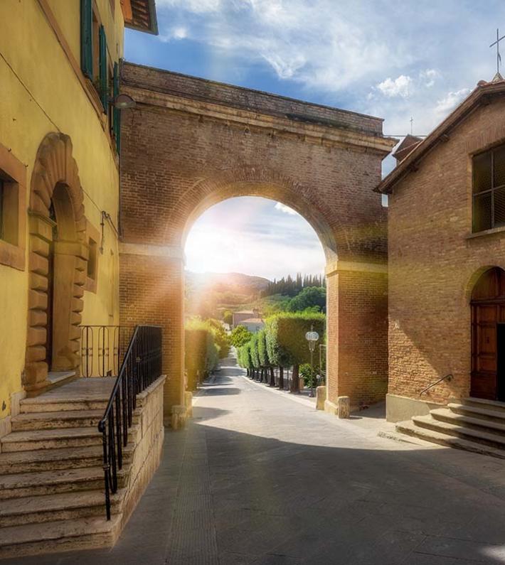 Strada soleggiata con arco antico e chiesa in mattoni.